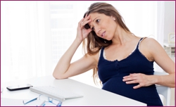 Tired brown haired pregnant woman sitting at the desk with hand on her belly and hand in hair while looking away.